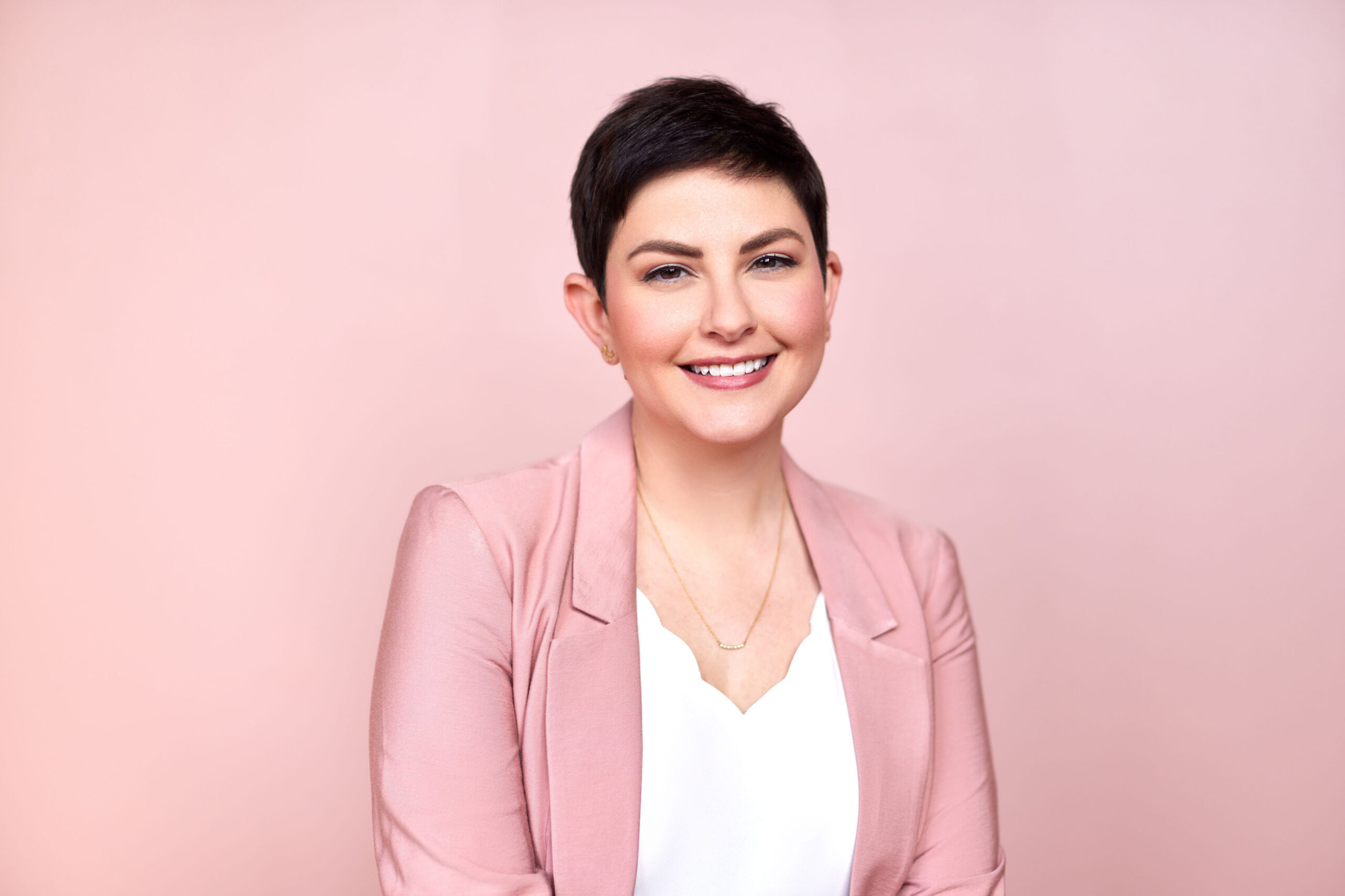 A woman with short, brown hair smiling at the camera. She is wearing a pink blazer and white shirt underneath.