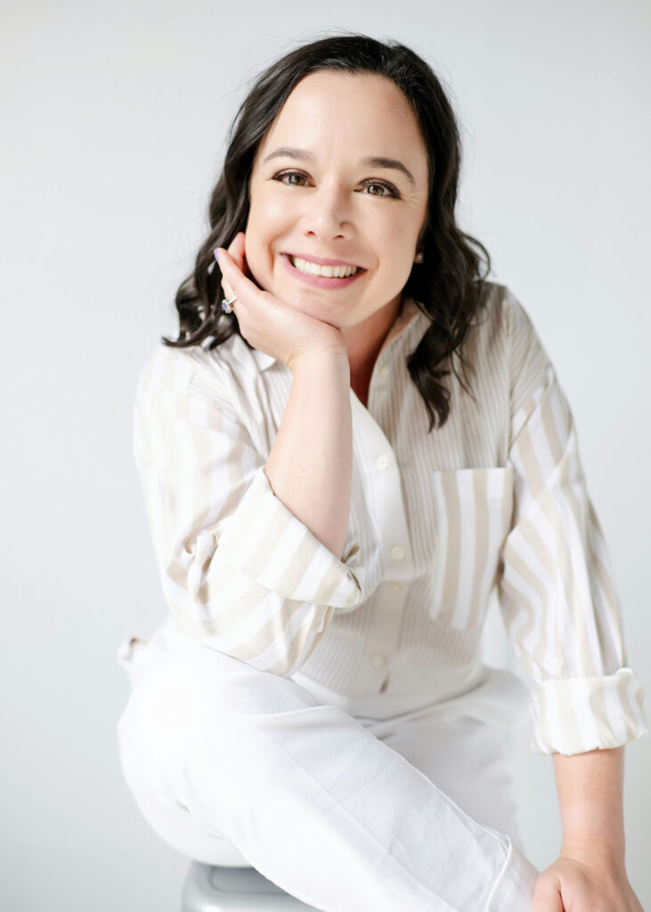 A brown-haired woman sitting on a stool wearing white pants and a striped button down shirt, smiling at the camera