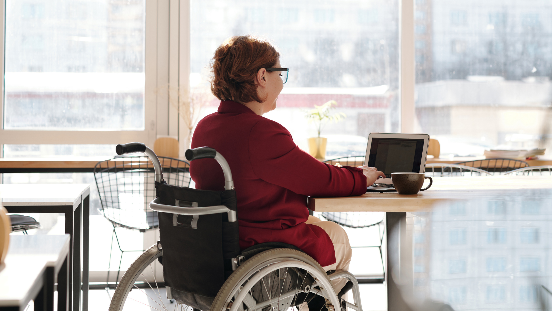 An image of a woman working at a desk wearing a maroon blazer, sitting in a wheelchair.